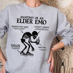 May include: A light gray sweatshirt with the text "ANATOMY OF AN ELDER EMO" and a black graphic of a skeleton. The graphic includes labeled body parts and a coffee cup. The brand "Briggs Design Apparel" is at the bottom.