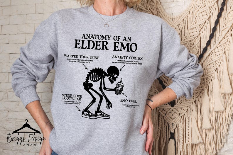 May include: A light gray sweatshirt with the text "ANATOMY OF AN ELDER EMO" and a black graphic of a skeleton. The graphic includes labeled body parts and a coffee cup. The brand "Briggs Design Apparel" is at the bottom.
