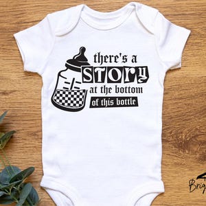 May include: White baby onesie with a black and white graphic of a baby bottle and the text "there's a STORY at the bottom of this bottle". The onesie has short sleeves and a round neckline. The Briggs Design Apparel logo is at the bottom.