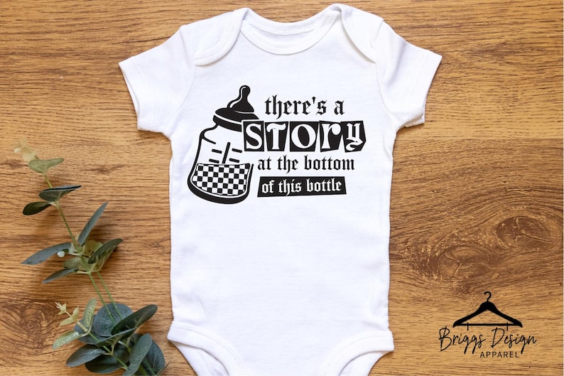 May include: White baby onesie with a black and white graphic of a baby bottle and the text "there's a STORY at the bottom of this bottle". The onesie has short sleeves and a round neckline. The Briggs Design Apparel logo is at the bottom.