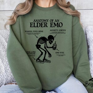 May include: Olive green sweatshirt with the text "ANATOMY OF AN ELDER EMO" and a skeleton graphic. The graphic includes labels for "Warped Tour Spine", "Anxiety Cortex", "Scene-Core Footwear", and "Emo Fuel".