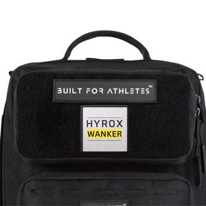 May include: Black backpack with a top handle and multiple compartments. The front features a rectangular patch with the text "BUILT FOR ATHLETES" above a smaller patch that reads "HYROX WANKER" in yellow text.