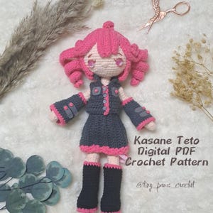 May include: A crochet doll with pink hair, a black and grey outfit, and pink boots. The doll is holding a small, pink flower. The text "Kasane Teto Digital PDF Crochet Pattern @tiny_paws_crochet" is written below the doll.