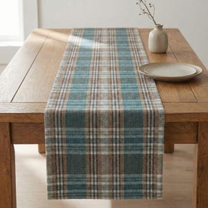May include: A plaid table runner in shades of teal, brown, and white, displayed on a wooden table. The runner features a classic check pattern, adding a touch of rustic charm to the dining setting. A small vase and plate are also on the table.