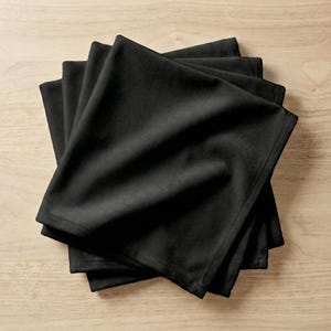 Può includere: Una pila di cinque tovaglioli di stoffa neri, ben piegati. I tovaglioli sono realizzati in un materiale morbido e opaco e sono disposti su una superficie di legno chiaro. Il colore scuro dei tovaglioli contrasta con lo sfondo chiaro.