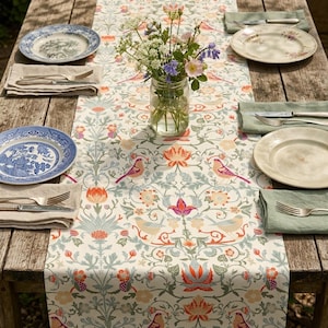 May include: A table runner featuring a repeating pattern of birds, orange and blue flowers, and green foliage on a cream background. A glass vase filled with wildflowers sits in the centre, surrounded by four place settings with plates and silverware.