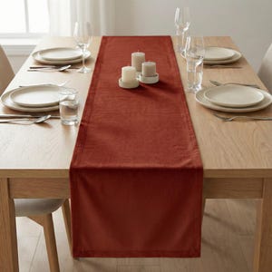 Könnte beinhalten: Ein Esstisch, der für eine Mahlzeit gedeckt ist. Ein rostfarbener Tischläufer ist auf dem hellen Holztisch zentriert. Weiße Teller, Besteck und Gläser sind für jeden Gedeck platziert. Drei weiße Kerzen stehen auf dem Läufer.