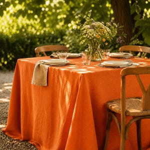 May include: An outdoor dining scene featuring a vibrant orange tablecloth draped over a wooden table. The table is set with white plates, clear glasses, and a vase of wildflowers. Wooden chairs surround the table, creating a warm and inviting atmosphere.
