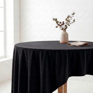 May include: A round black velvet tablecloth on a wooden table. A small vase with greenery and a folded napkin are on the table. The room has a white wall and a window.