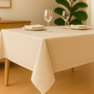 May include: A beige rectangular tablecloth covers a wooden table. Two wine glasses, plates, and napkins are set on the table. The table has wooden legs. A plant is in the background.