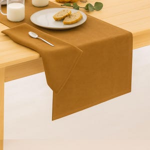 May include: A table setting featuring a mustard-colored table runner and matching napkin. A glass of milk, a bottle of milk, and a plate with bread slices are on the wooden table. A silver spoon rests on the napkin.