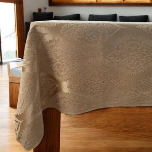 Könnte beinhalten: Eine beige Tischdecke mit Damastmuster. Die Tischdecke ist über einen Holztisch gelegt.