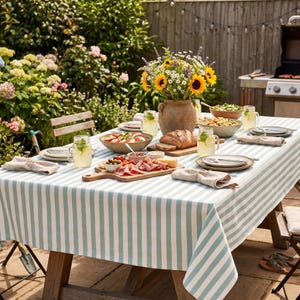 Può includere: Un tavolo da picnic apparecchiato per un raduno all'aperto, con una tovaglia a righe bianche e azzurre. Il tavolo è imbandito con cibo, bevande e un vaso di girasoli. La scena è ambientata in un giardino con un barbecue sullo sfondo.