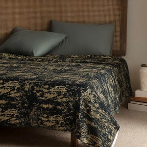 Peut inclure: Un lit avec une couverture sombre à motif abstrait doré et noir. Deux oreillers gris reposent contre une tête de lit en osier. Un vase blanc et des livres sont visibles sur le côté droit du lit, suggérant une chambre.