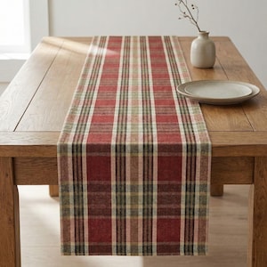 May include: A red and green plaid table runner on a wooden table. The runner has a traditional plaid design in red, green, and beige. A small vase with a sprig and a plate are on the table.