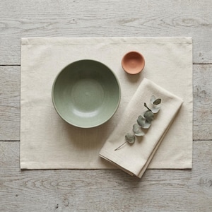 Puede incluir: Una composición de naturaleza muerta con un cuenco de cerámica verde claro, un pequeño cuenco de terracota y una servilleta de lino beige doblada con eucalipto. Los artículos están sobre un mantel individual beige, sobre una superficie de vetas de madera clara.