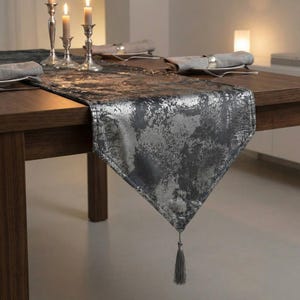 May include: A dark gray table runner with a silver metallic pattern and a tassel. The runner is draped over a wooden table, with lit candles and silverware visible in the background. The runner measures approximately 12 inches wide.