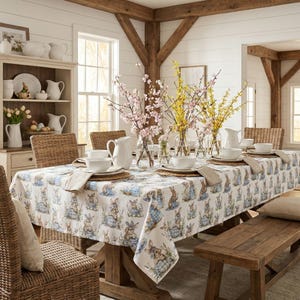 Puede incluir: Una mesa de comedor preparada para una celebración primaveral. La mesa está cubierta con un mantel blanco con un estampado de conejos. La vajilla blanca, las servilletas y los centros de mesa florales completan el conjunto. Sillas de mimbre y un banco de madera rodean la mesa.