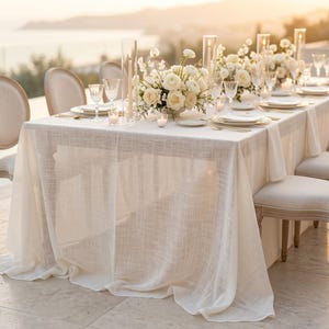 May include: A beautifully set table for an outdoor event. The table is covered with a white linen tablecloth and set with white plates, gold silverware, and crystal glassware. Centerpieces of white roses and candles add to the elegant ambiance.