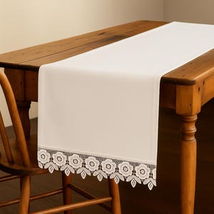 Może przedstawiać: Kremowy bieżnik na stół z delikatnym kwiatowym koronkowym wykończeniem. Bieżnik jest ułożony na drewnianym stole, prezentując swoją gładką fakturę i elegancki design. Koronkowe wykończenie dodaje elegancji nakryciu stołu.