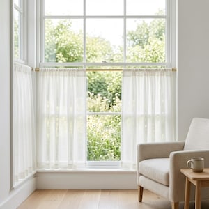 May include: A set of sheer, off-white cafe curtains hanging on a thin, horizontal brass rod. The curtains feature a simple rod pocket header and a straight hem, allowing soft light to filter through the fabric into the room interior.