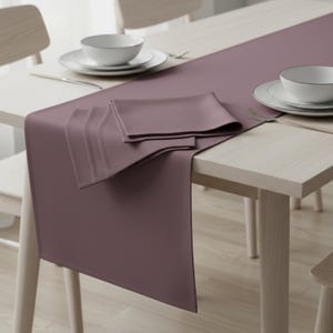 Könnte beinhalten: Ein mauvefarbener Tischläufer mit passenden Servietten und weißem Geschirr. Der Läufer ist über einem hellen Holztisch drapiert und schafft eine schlichte, elegante Essumgebung. Der Tisch ist mit Tellern, Schalen und Besteck gedeckt.