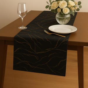 Puede incluir: Camino de mesa negro con un patrón de mármol dorado, sobre una mesa de madera. Una copa de vino, un plato blanco, un tenedor dorado y un jarrón con rosas blancas completan la mesa. El camino de mesa añade elegancia.