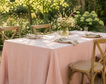 May include: A pale pink linen tablecloth on a wooden table, set for an outdoor meal. Plates, napkins, and glasses are arranged on the table, with a vase of flowers in the centre. Wooden chairs surround the table, set against a natural backdrop.