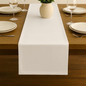 Peut inclure: Une table à manger dressée avec un chemin de table blanc, des assiettes, des verres à vin et des couverts. Un vase de marguerites blanches est au centre. La table est en bois foncé, créant une atmosphère fraîche et accueillante.