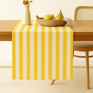 May include: A yellow and white striped table runner is draped over a wooden table. A woven basket holds two green pears, and a cream-colored vase holds dried branches. A wooden chair with a woven back is partially visible.