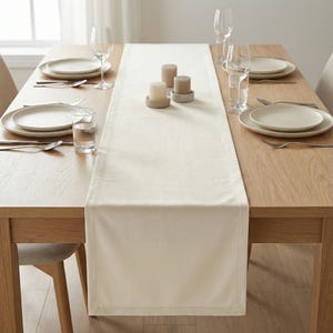 May include: A dining table set for a meal. The wooden table is set with cream-colored plates, silverware, and wine glasses. A cream-colored table runner runs down the center, with candles in the middle. The table is set for four people.