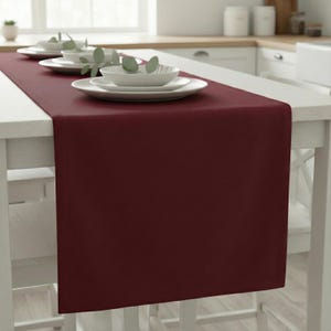 Puede incluir: Un camino de mesa burdeos sobre una mesa blanca, con platos y cuencos blancos. El camino de mesa es de color liso y parece estar hecho de una tela suave. La mesa está puesta en una cocina.