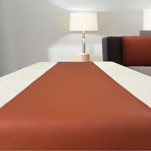 Könnte beinhalten: Ein rostfarbener Tischläufer auf einem weißen Tisch. Der Läufer ist aus einem glatten, seidenartigen Material gefertigt.