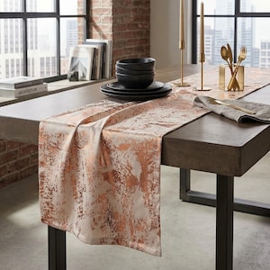 May include: A dining table is set with black bowls and plates, gold cutlery, and a cream-colored table runner with a copper-colored abstract design. Two gold candle holders and candles are on the table. The table is set in front of a window.