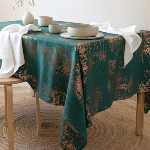 Può includere: Una tovaglia verde con un motivo astratto color rame copre un tavolo di legno. Tovaglioli di lino bianco e stoviglie in ceramica, tra cui una ciotola, un piatto e tazze, sono disposti sul tavolo. È presente anche un vaso con steli secchi.