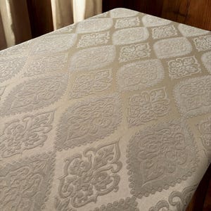 Puede incluir: Primer plano de una tela beige con un patr&oacute;n en relieve de diamantes blancos y dise&ntilde;os florales. La tela tiene un tacto suave y texturizado.