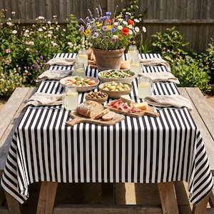 May include: A garden picnic scene with a black and white striped tablecloth. The table is laden with food, drinks, and a pot of flowers. Plates, glasses, and napkins are arranged for a meal. The table is made of wood.