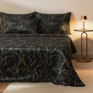 Peut inclure: Parure de lit en satin noir avec un motif abstrait doré. L'ensemble comprend un drap-housse, un drap plat et deux taies d'oreiller. Le lit est fait avec les draps et les taies d'oreiller. Une petite lampe dorée est posée sur une table de chevet.
