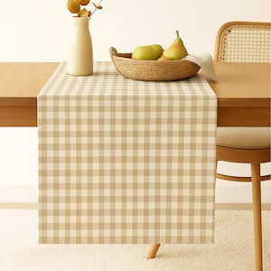 Könnte beinhalten: Ein beige-weiß karierter Tischläufer auf einem Holztisch. Ein geflochtener Korb enthält grüne Birnen, und eine weiße Vase enthält getrocknete Blätter. Ein Holzstuhl ist teilweise sichtbar.