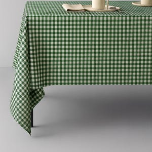 Op de afbeelding: Een groen-wit geblokt tafelkleed bedekt een tafel, met een deel van de stof dat over de rand hangt. Twee beige mokken en een gevouwen servet liggen op de tafel. Het tafelkleed heeft een klassiek ruitpatroon.