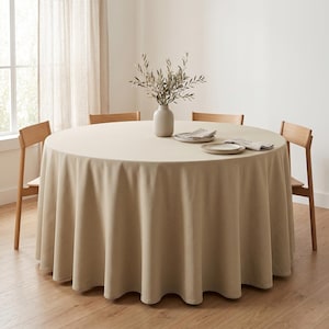 May include: A round dining table covered with a solid beige tablecloth, surrounded by four light wood chairs. A matte beige vase containing green olive branches sits in the center, accompanied by two small plates topped with folded beige napkins.