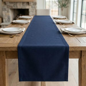 May include: A navy blue table runner is centered on a light brown wooden dining table. White plates, gold forks, and beige napkins are set for a meal. A stone fireplace and window are visible in the background.