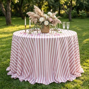 Könnte beinhalten: Ein runder Tisch mit einer rosa-weiß gestreiften Tischdecke. Der Tisch ist mit einem Blumenarrangement, Kerzen und Sektgläsern dekoriert. Die Szene spielt sich im Freien auf einer Rasenfläche ab.