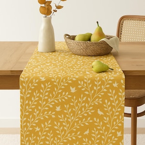 May include: Mustard yellow table runner with a white leaf and bird pattern. A white vase with dried branches and a woven basket with pears sit on the table. The table is made of wood, and a chair is partially visible.