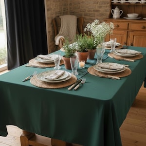 Könnte beinhalten: Ein Esstisch, der für eine Mahlzeit gedeckt ist und mit einer dunkelgrünen Tischdecke bedeckt ist. Jedes Gedeck umfasst einen Teller, eine Serviette und Besteck auf einem gewebten Platzset. Kleine Topfpflanzen und Blumen schmücken den Tisch. Der Tisch ist aus Holz.