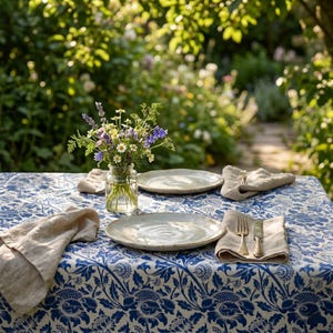 Può includere: Un tavolo apparecchiato per mangiare all'aperto con una tovaglia a fiori blu e bianca. Il tavolo è apparecchiato con piatti, tovaglioli e posate. Un piccolo vaso di vetro contiene un bouquet di fiori di campo. Lo sfondo è un giardino sfocato.