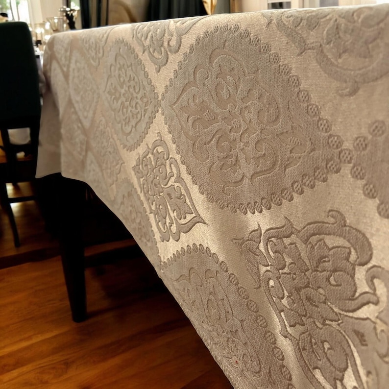 Puede incluir: Primer plano de un mantel beige con un patr&oacute;n de damasco. El mantel est&aacute; hecho de una tela suave y texturizada con un brillo sutil.