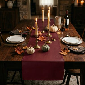 May include: A wooden dining table set for a fall-themed dinner. The table is adorned with a burgundy runner, lit candles in brass holders, pumpkins, and fall leaves. Wine glasses and plates are set for guests.