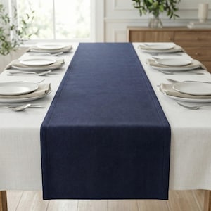 Könnte beinhalten: Ein marineblauer Tischläufer liegt mittig auf einer weißen Tischdecke, mit Gedecken auf beiden Seiten. Weiße Teller, Besteck und beige Servietten sind arrangiert. Der Essbereich ist hell und einladend.