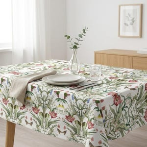 May include: A white tablecloth featuring a repeating botanical pattern of red tulips, green foliage, and small butterflies. A beige linen napkin is folded on the left, with a stack of white ceramic plates and a clear glass vase in the centre.